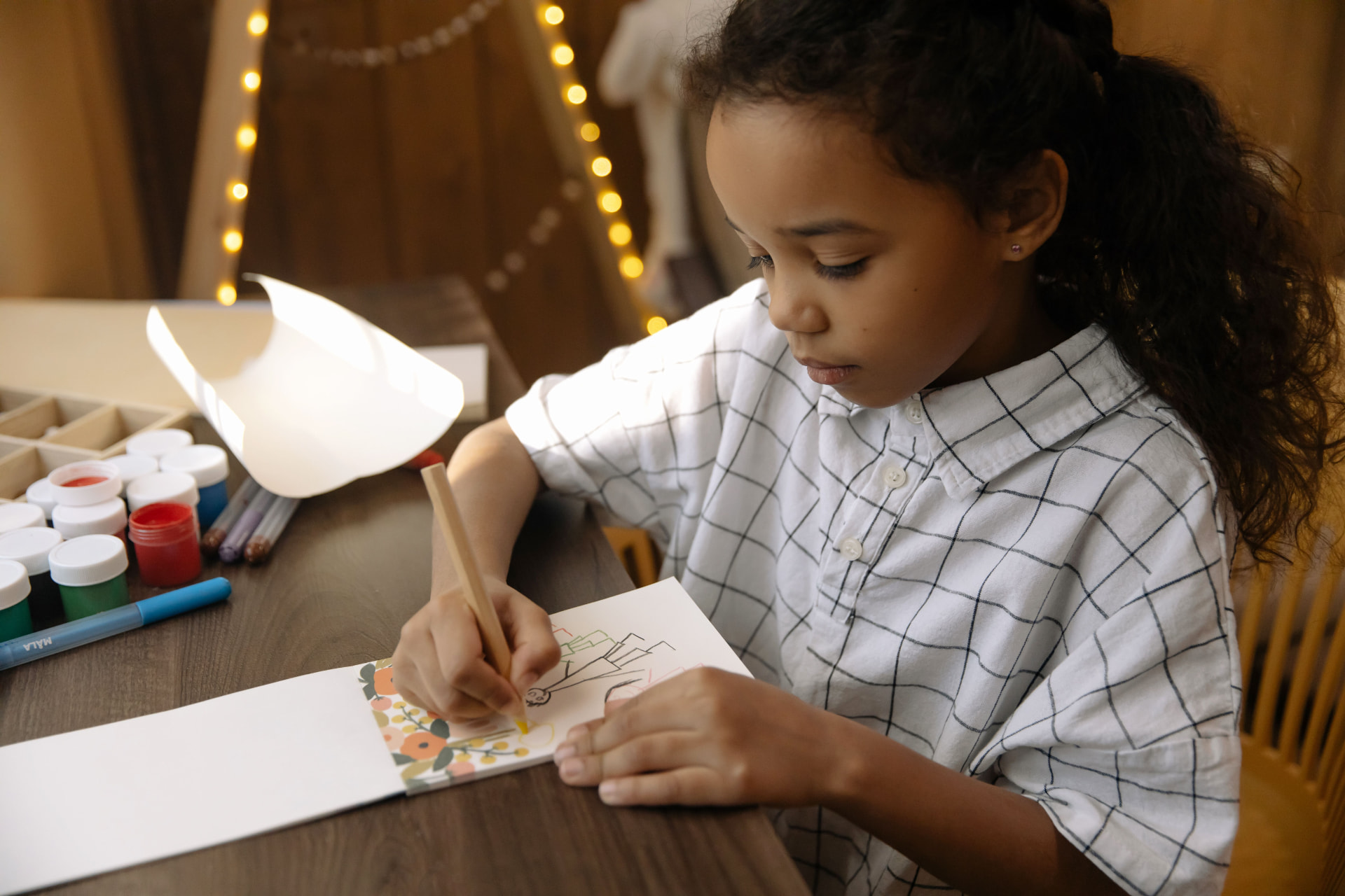 A girl drawing a holiday card at a craft table under string lights